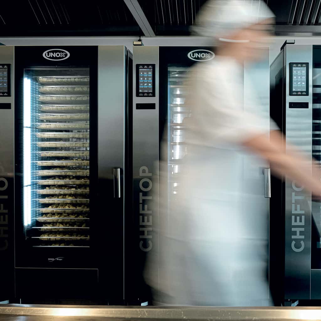 Kitchen with two tall combi trolley ovens next to each other, a chef with a white hat walks past the camera pushing a trolley, causing motion blur.