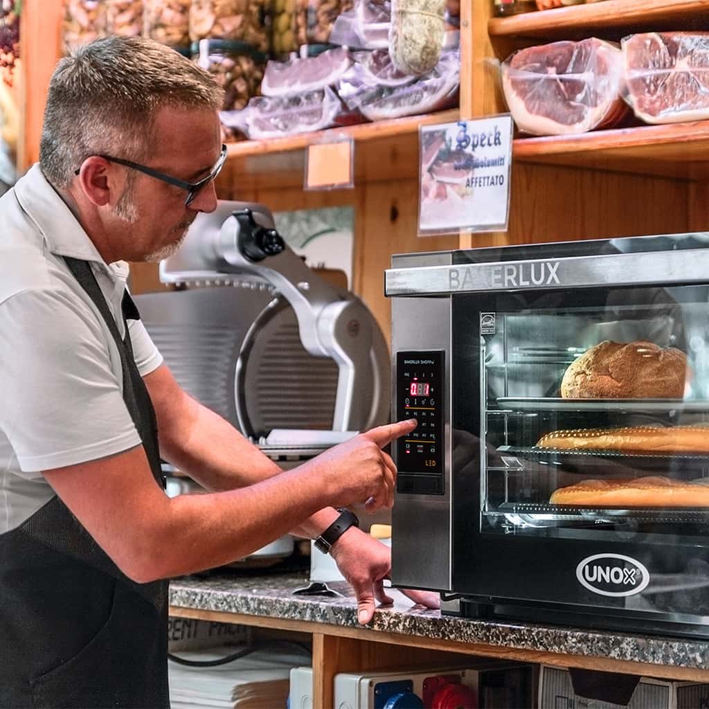 Man in deli pushing button on Unox Bakerlux baking oven, with bread baking inside oven, meat slicer and different meats can be seen.