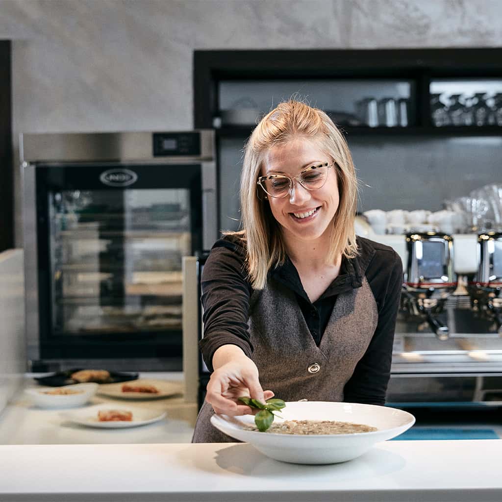 Smiling woman with glasses adding herbs to a dish of risotto, coffee machines, dishes of food and Unox Evereo can be seen behind her.
