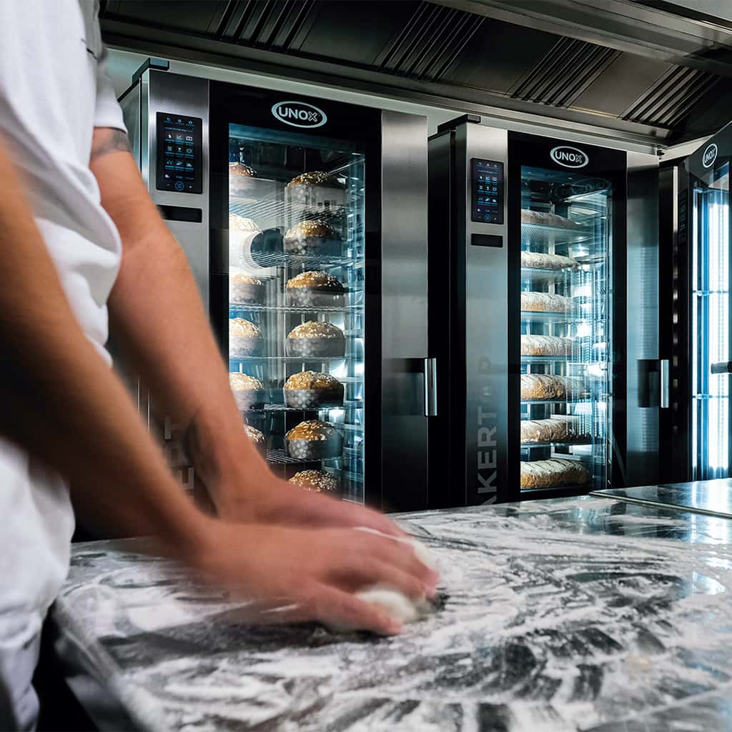 Man's hands rolling out dough on a flour-topped counter, Unox bakertop tall trolley ovens filled with panettone and bread can be seen in background.