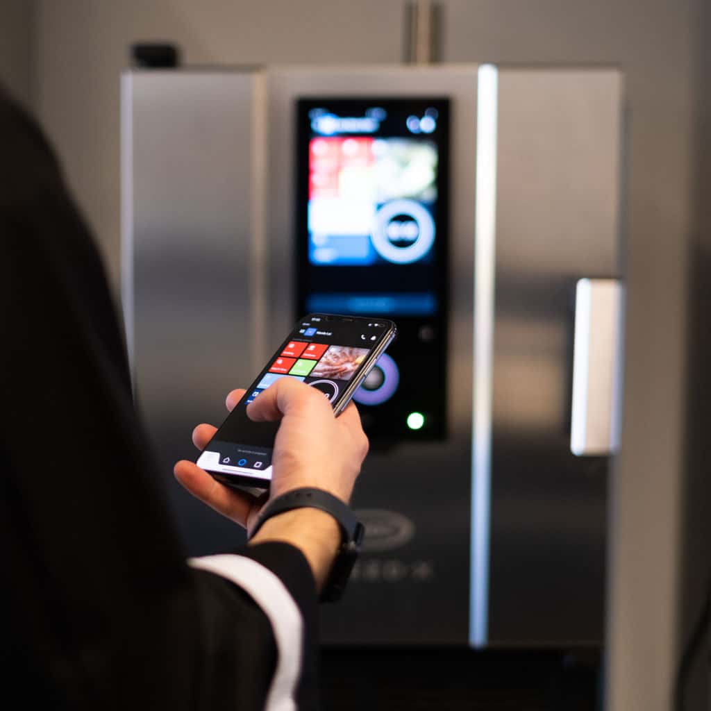 Man in black chef jacket holding a smart-phone pressing some buttons, facing towards an unox speed-x speed oven, controlling the oven with his phone.