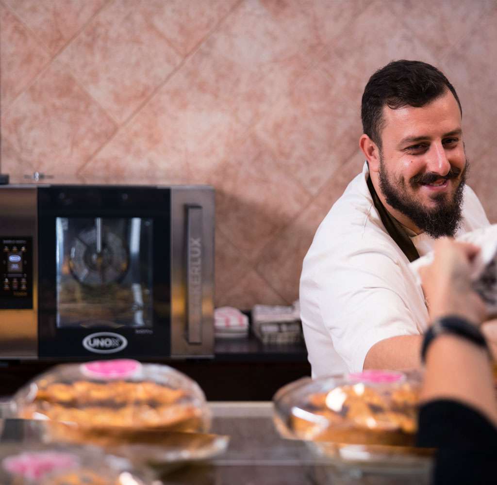 A male operator serves a customer behind the counter of a bar, with a small Unox commercial oven visible in the background