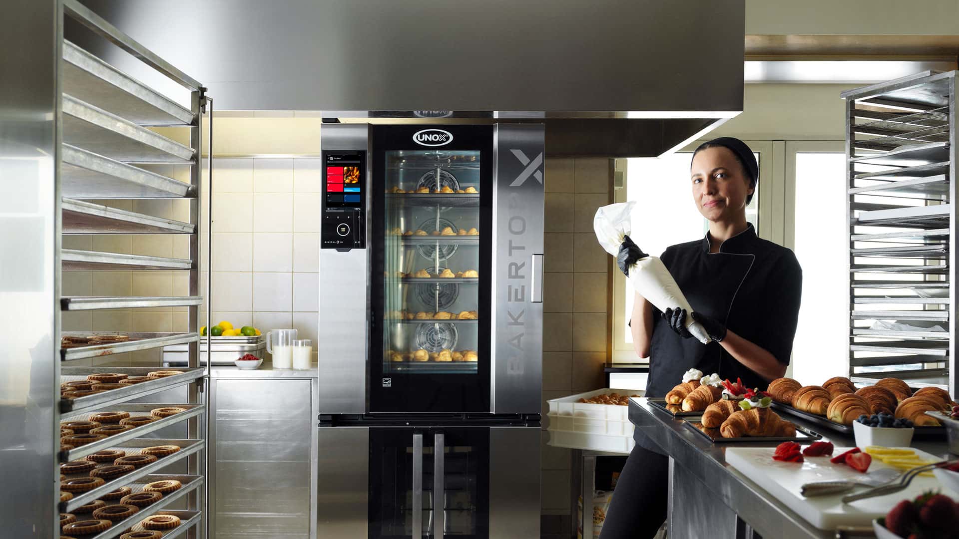 A baker in a kitchen, standing near an Unox combi oven with pastries inside, holds a piping bag. Pastries are displayed on a nearby countertop.