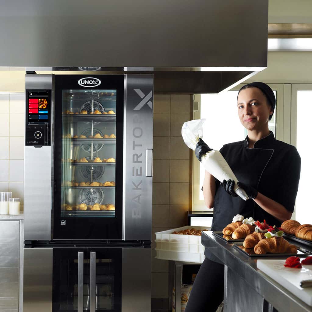 A baker in a kitchen, standing near an Unox combi oven with pastries inside, holds a piping bag. Pastries are displayed on a nearby countertop.