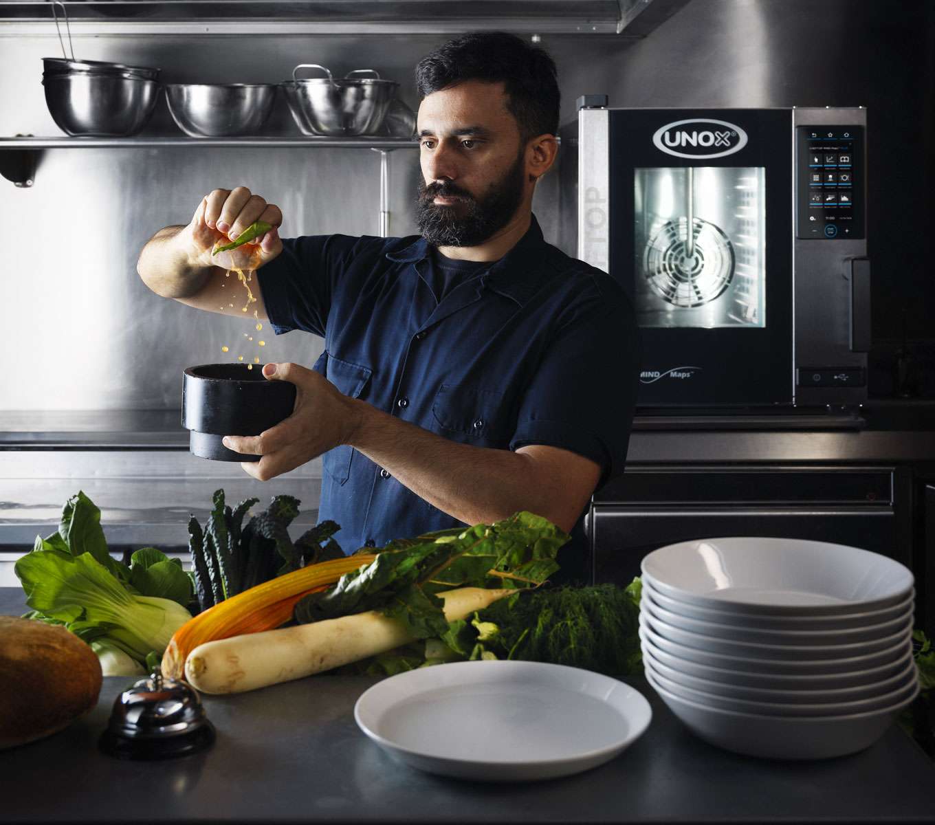 A chef works on a professional kitchen counter, preparing food with dishes and ingredients visible, while pots and a Unox small commercial oven are in the background