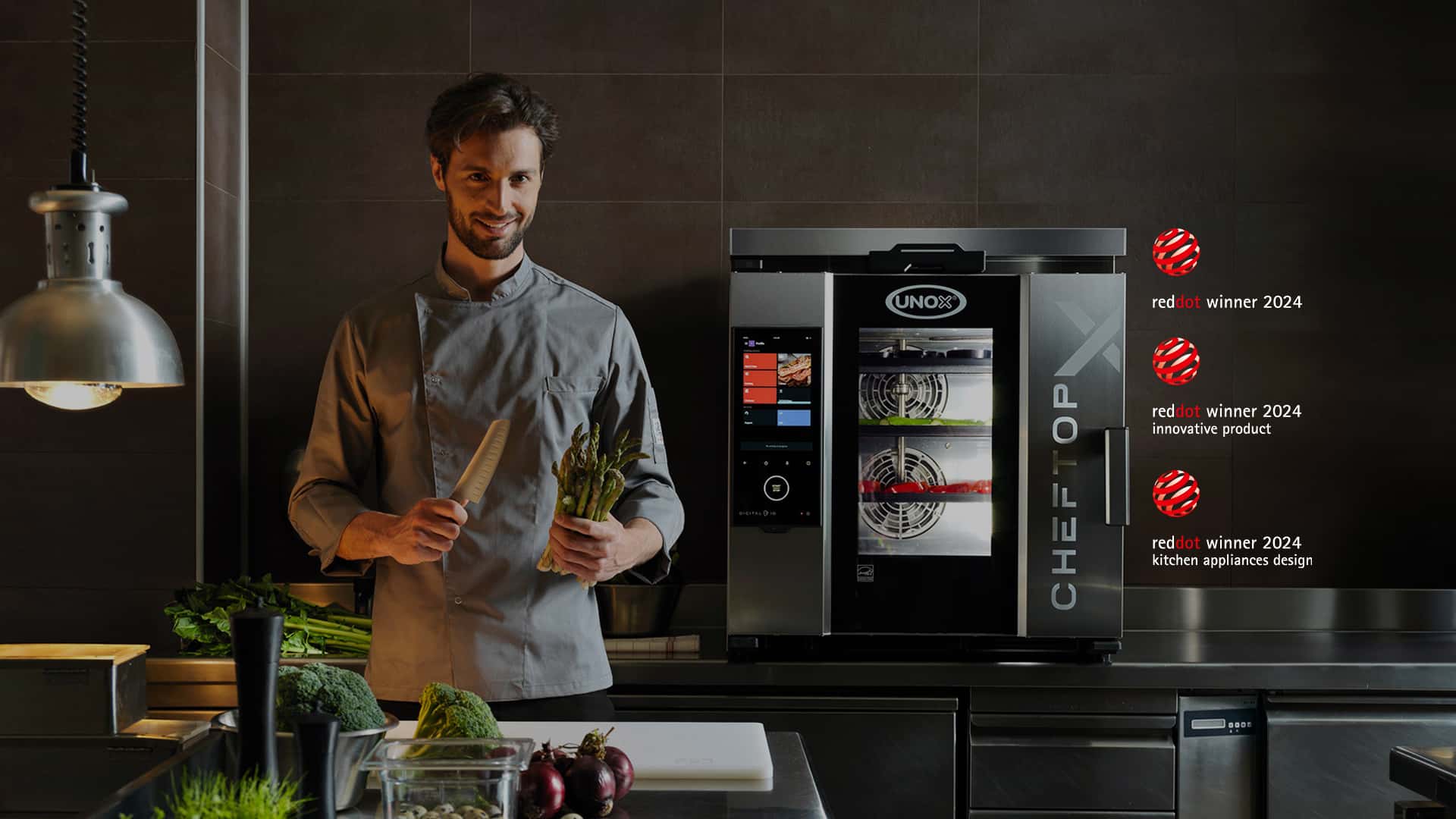 Smiling male Chef standing in a kitchen holding a knife and asparagus, next to an Unox combi oven with three Red Dot 2024 winner badges.