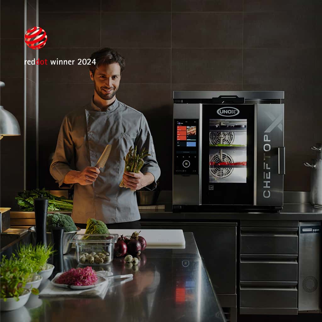 Smiling male Chef standing in a kitchen holding a knife and asparagus, next to an Unox combi oven with three Red Dot 2024 winner badges.