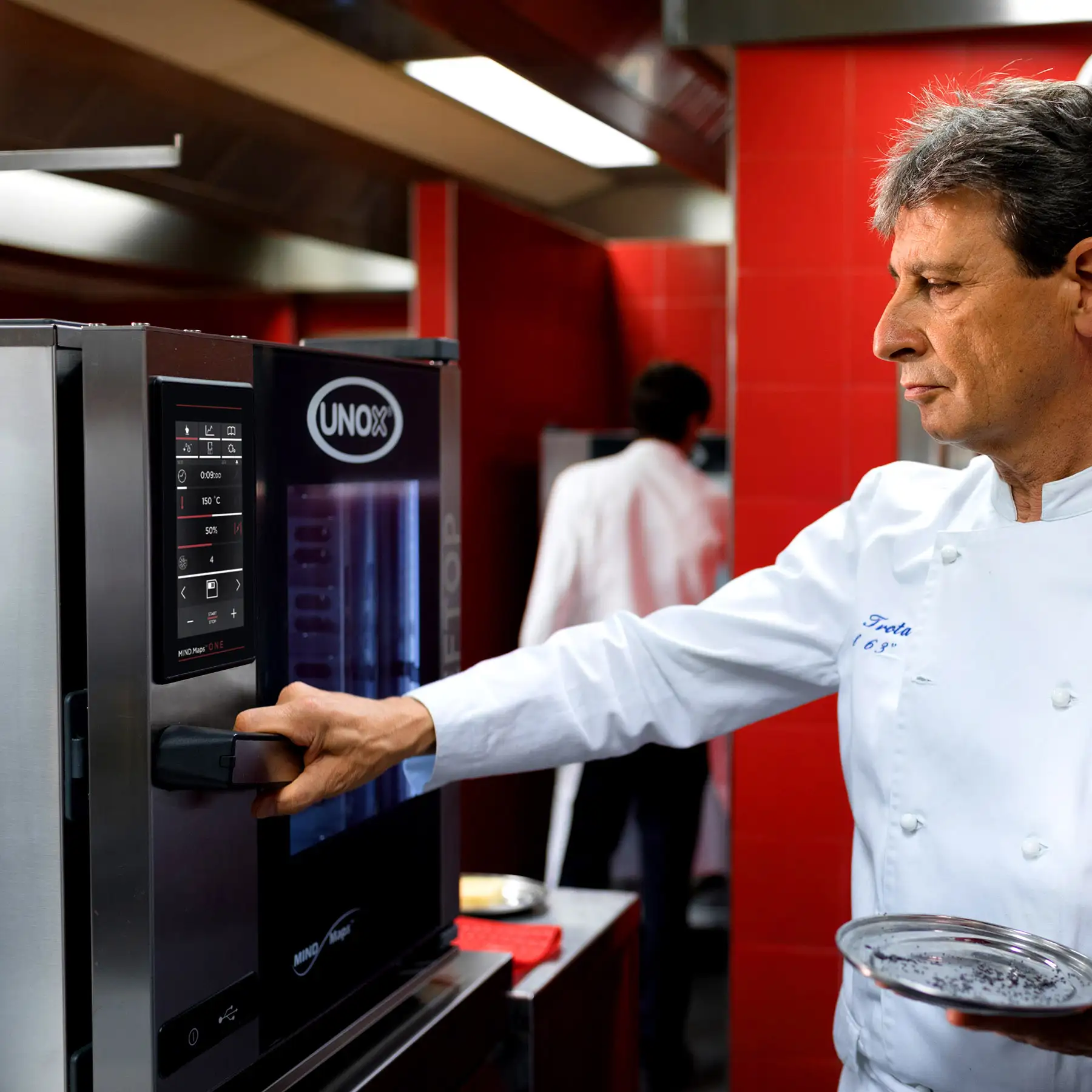 Male chef in white jacket opening door of an Unox combi oven, holding silver plate in his other hand.