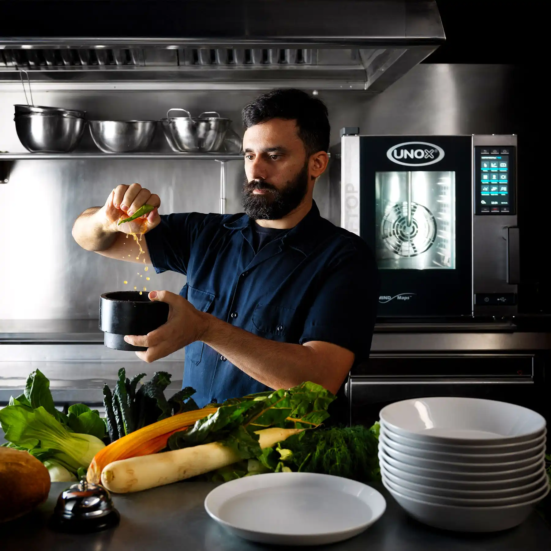 Male chef in kitchen stood in front of an Unox combi oven, holding a bowl and squeezing lime juice into the bowl, vegetables and plates on counter.