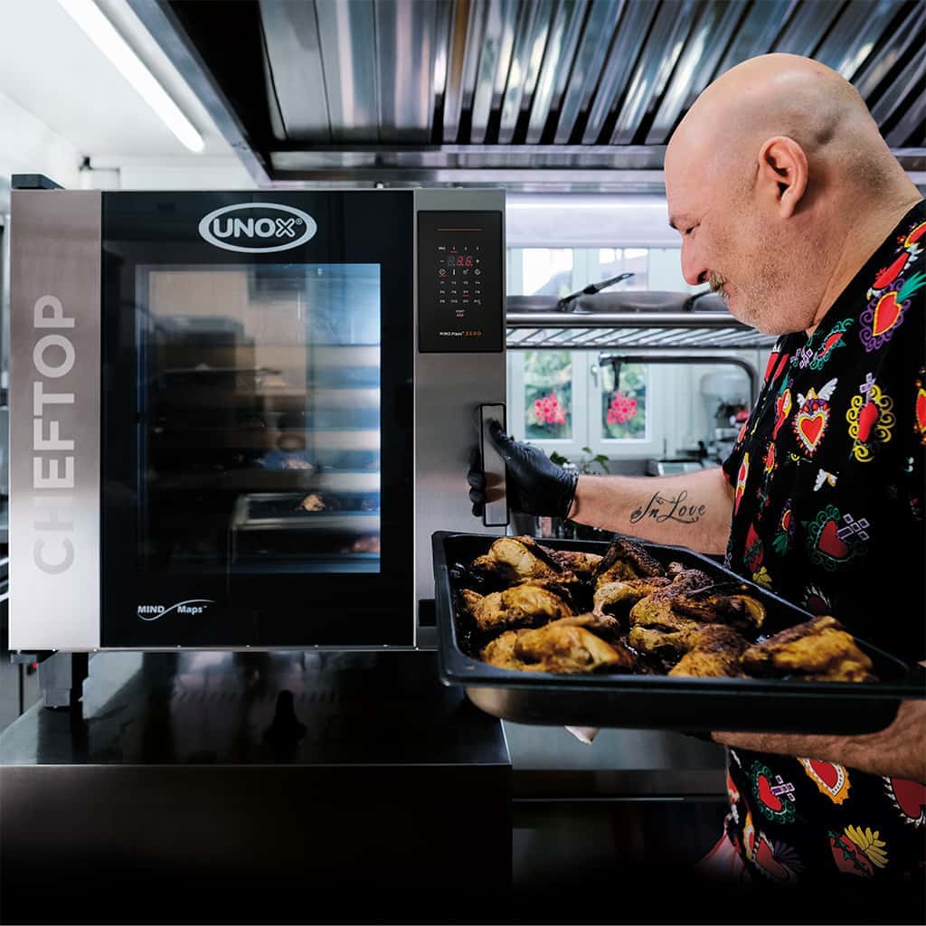 Man opening the door to an Unox combi oven with one hand, whilst holding a tray of cooked chicken legs in the other hand.