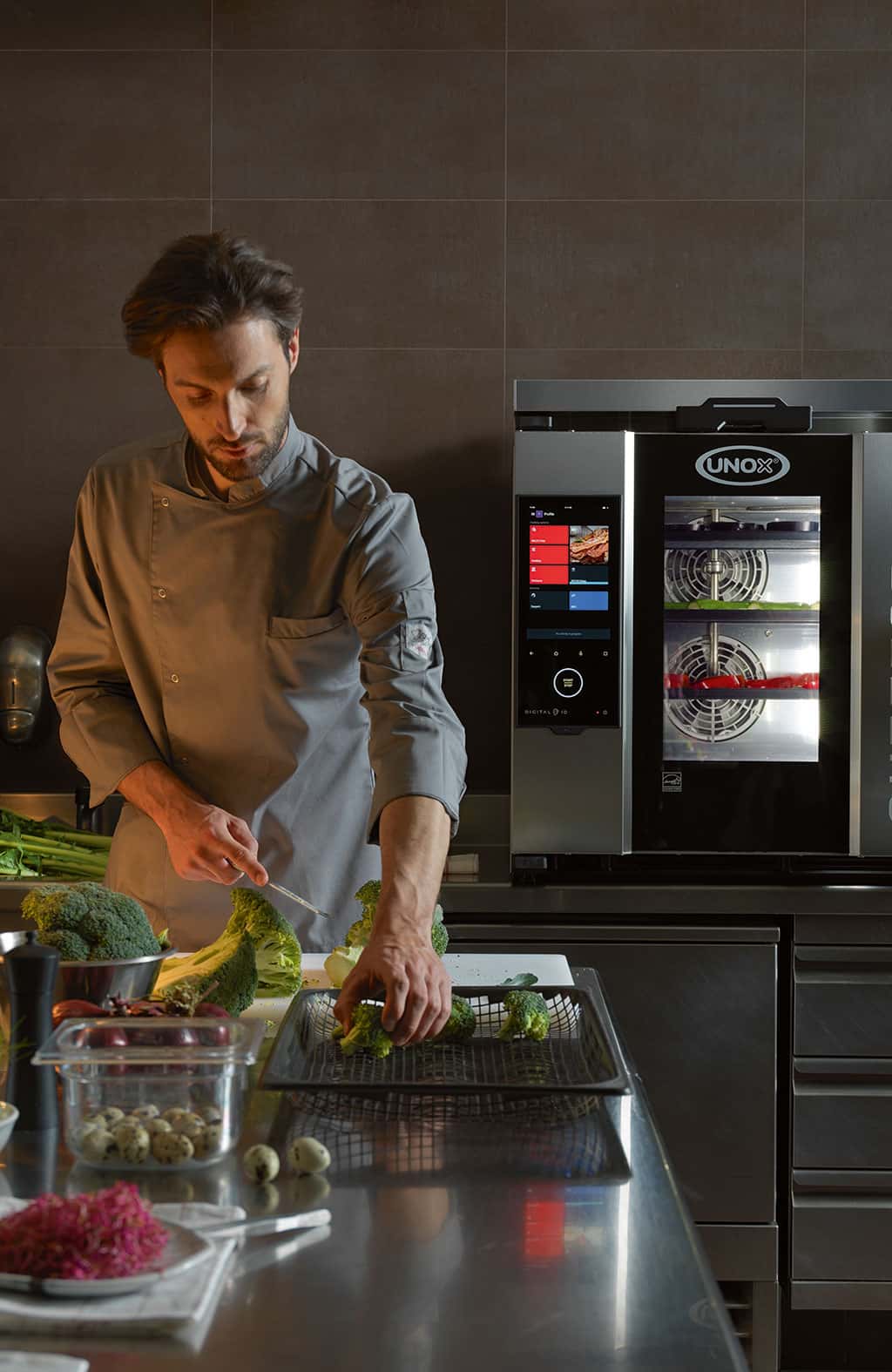 Male Chef standing in a kitchen holding a knife and broccoli, next to an Unox combi oven.