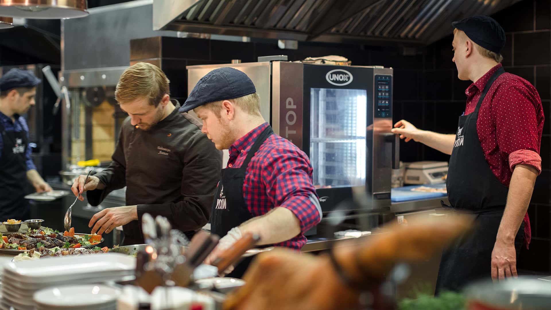 Team of male chefs working in a professional kitchen, preparing dishes with a Unox commercial combi oven in the background.