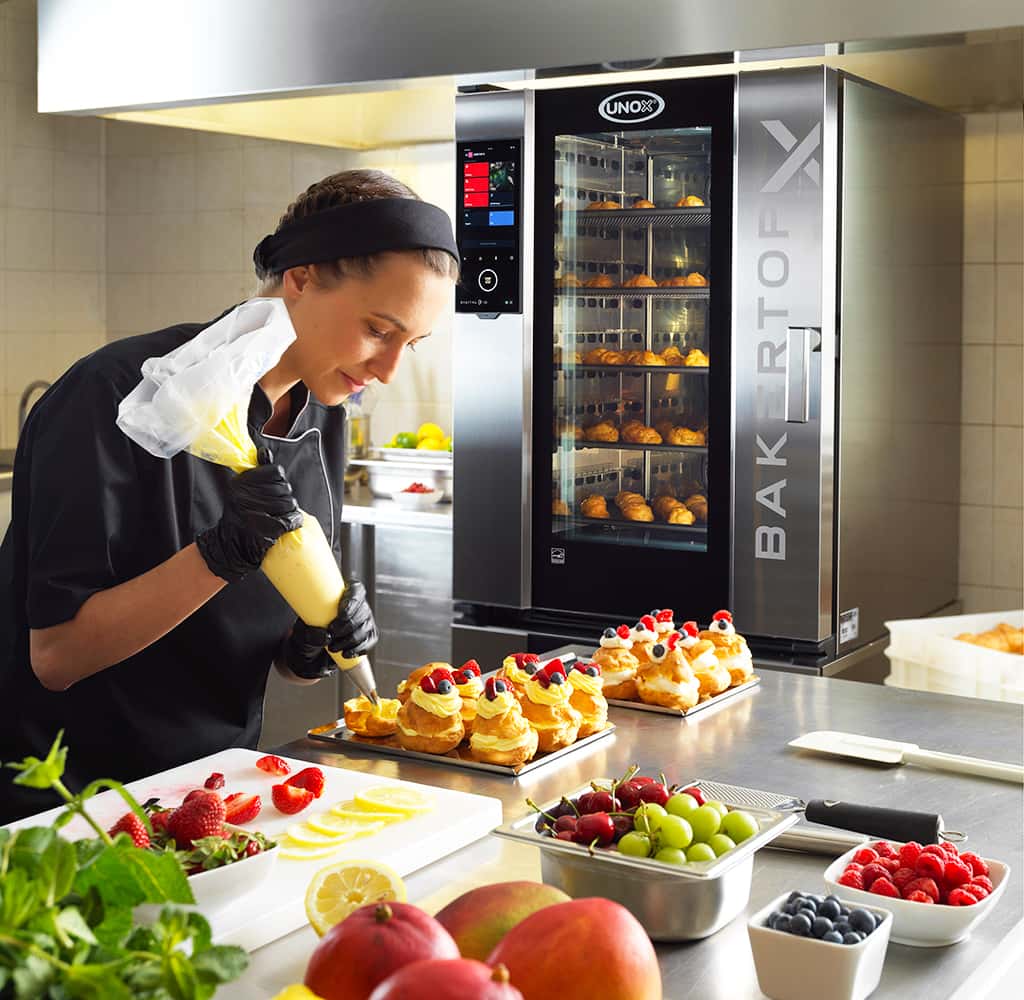 Female chef holding piping bag, adding cream to pastries, an assortment of berries are on the counter and an Unox baking oven filled with pastries.
