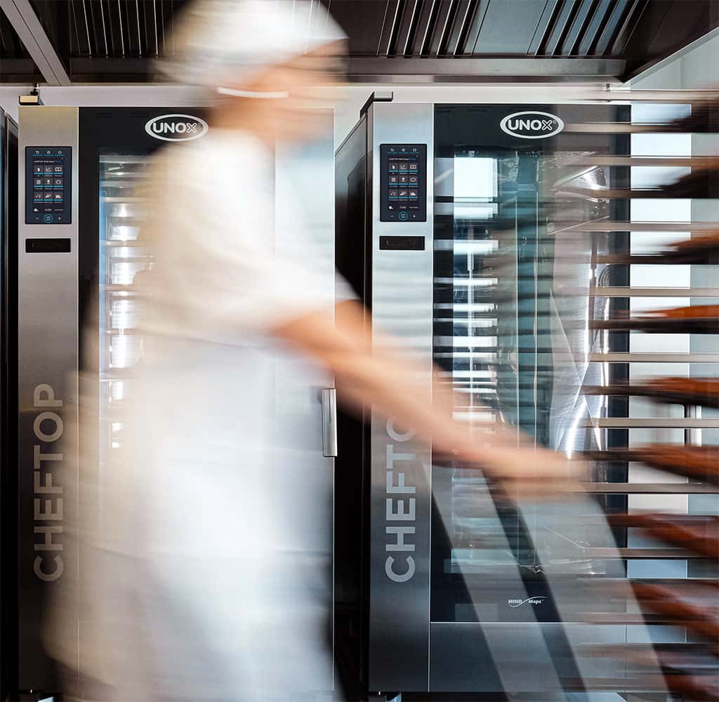 Kitchen with two tall combi trolley ovens next to each other, a chef with a white hat walks past the camera pushing a trolley, causing motion blur.
