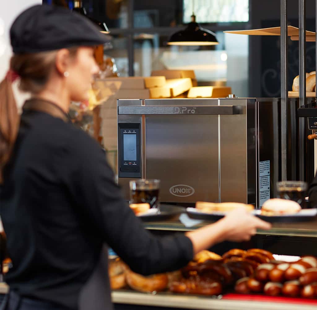 Unox speed oven on a counter in a coffee shop, a woman wearing a black cap and an apron is holding an assortment of sandwiches.