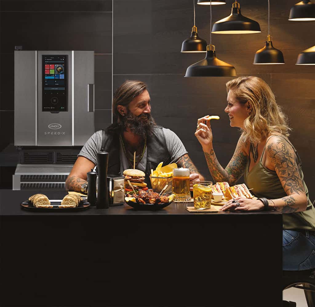 A smiling man and woman with tattoos sit at a table with burgers, chips, sandwich and other food, beer, a cocktail and an Unox speed oven behind them.