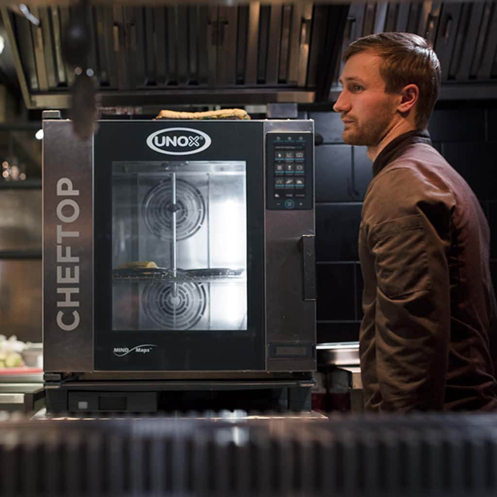Male chef standing beside a Unox CHEFTOP MINDMaps commercial combi oven in a professional kitchen setting with food inside.