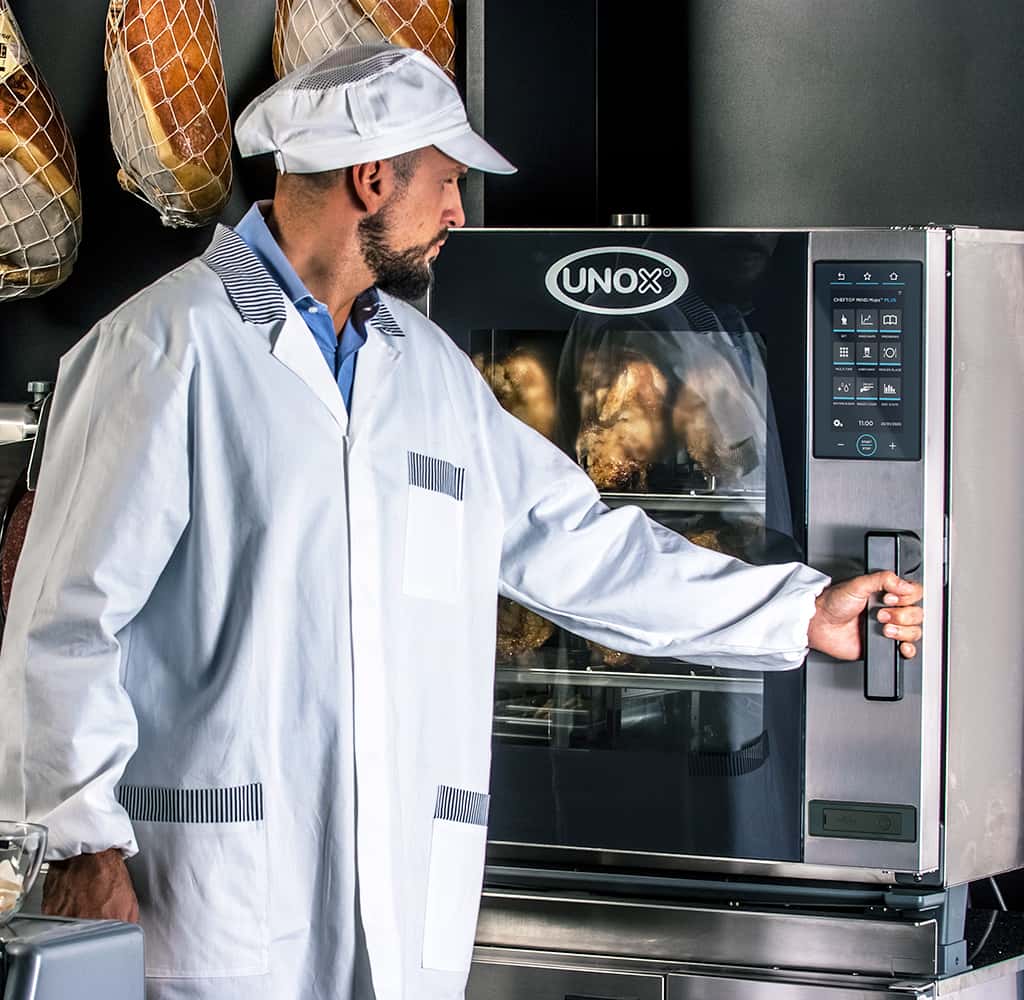 Man in a deli or butchers wearing a white hat, hams hang in the background, opening the door of an Unox combi oven filled with roasted chickens.