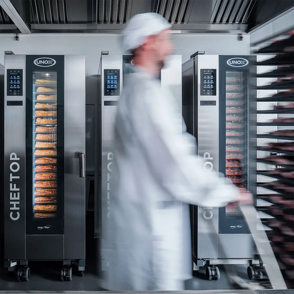 Kitchen with three tall combi trolley ovens next to each other, a chef with a white hat walks past the camera pushing a trolley, causing motion blur.