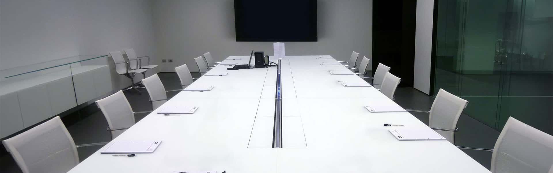 An office boardroom with a long white table in the middle, lined with white office chairs, white pads of paper and pens placed at each seat.