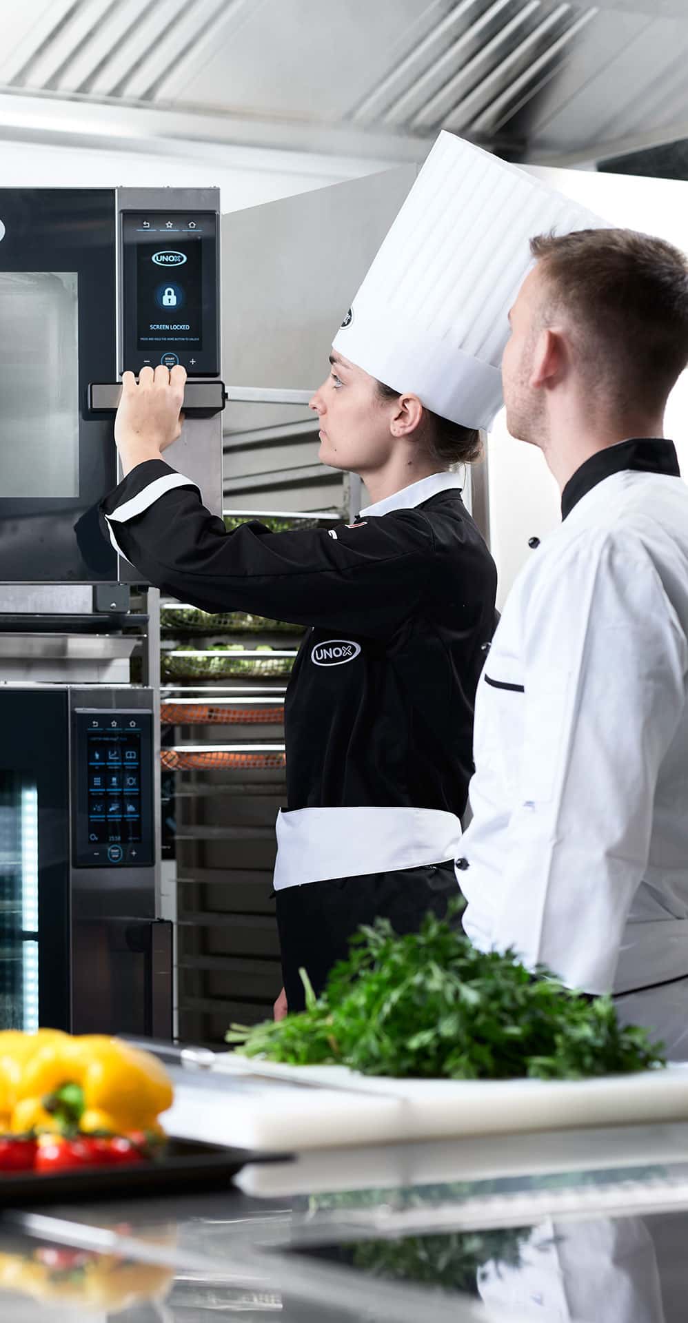 Female chef opening an unox combi oven door, male chef stands next to her in the kitchen, vegetables and herbs are on a chopping board on the counter.