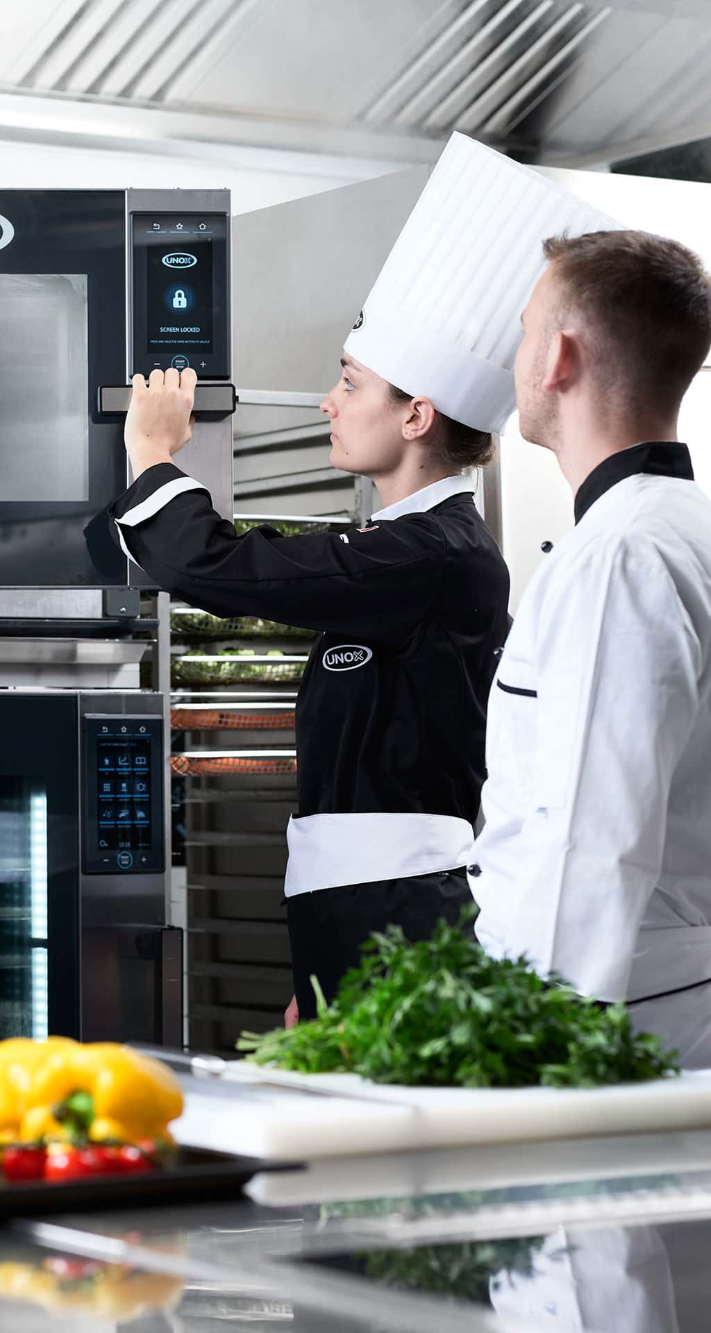 Female chef opening an unox combi oven door, male chef stands next to her in the kitchen, vegetables and herbs are on a chopping board on the counter.