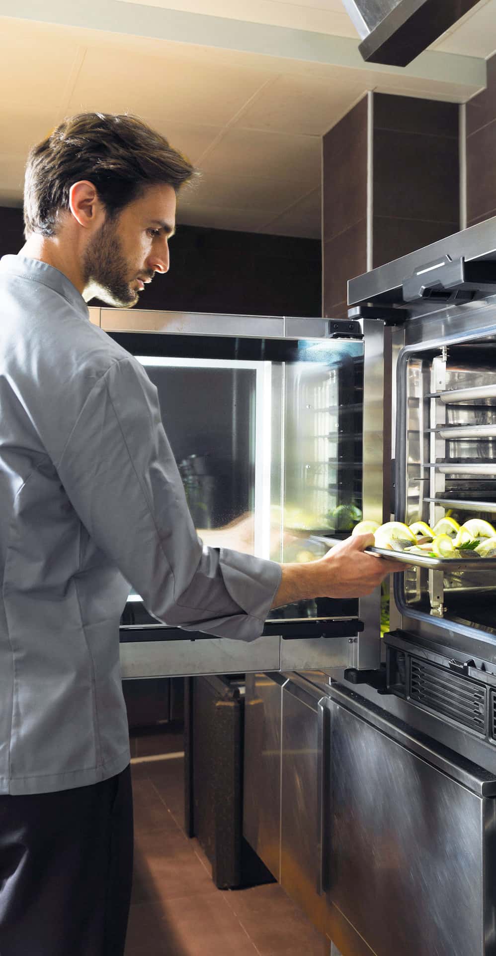 Male chef in kitchen opening comi oven door and inserting tray with fish and lemon into the oven.