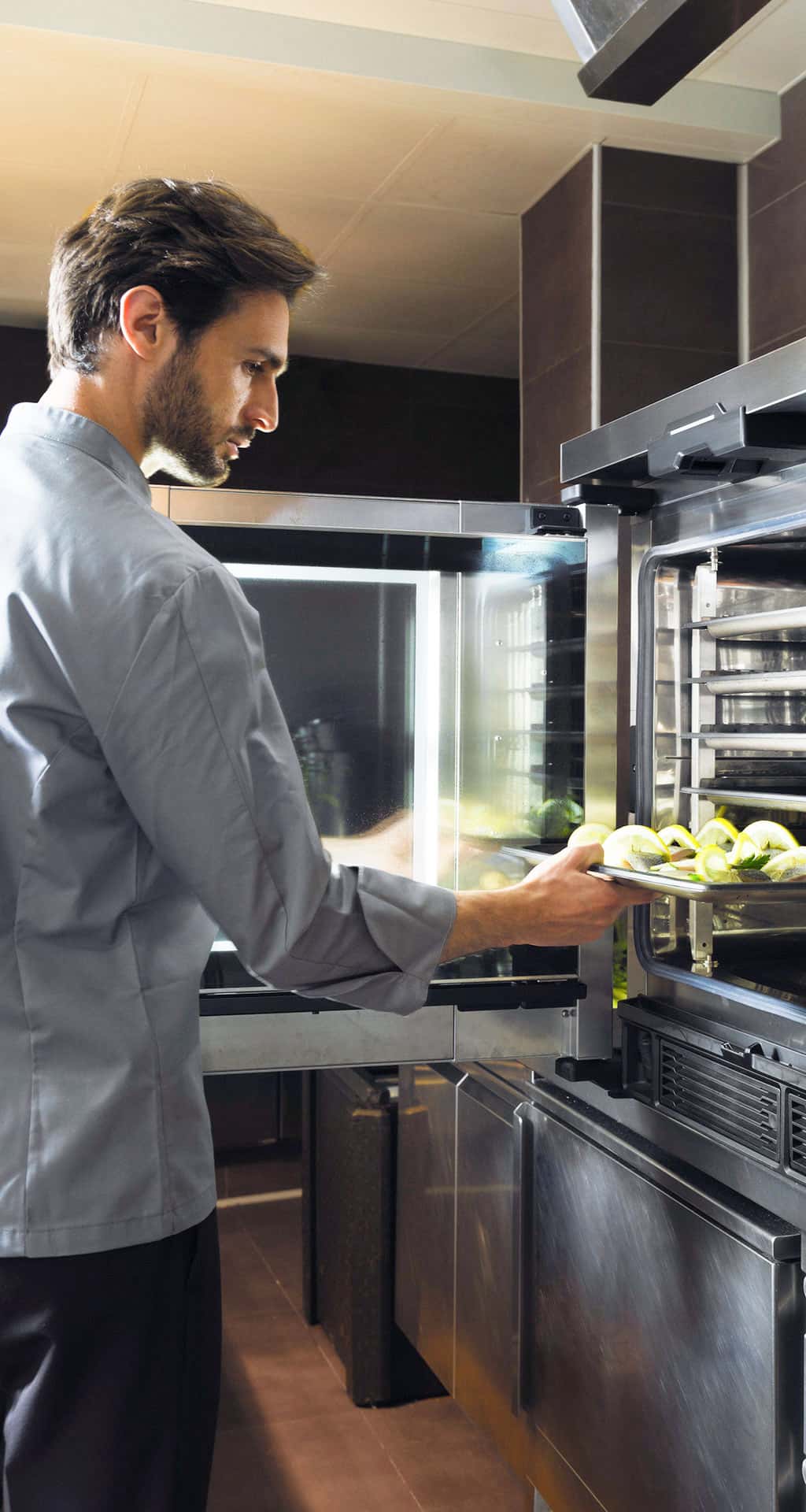 Male chef in kitchen opening comi oven door and inserting tray with fish and lemon into the oven.