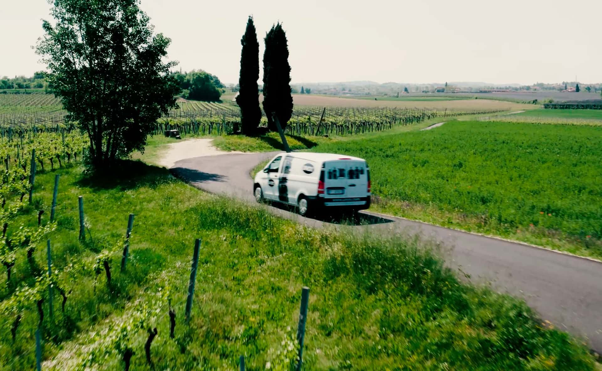 White van with Unox logo and images printed on drives down a road surrounded by green fields and trees.