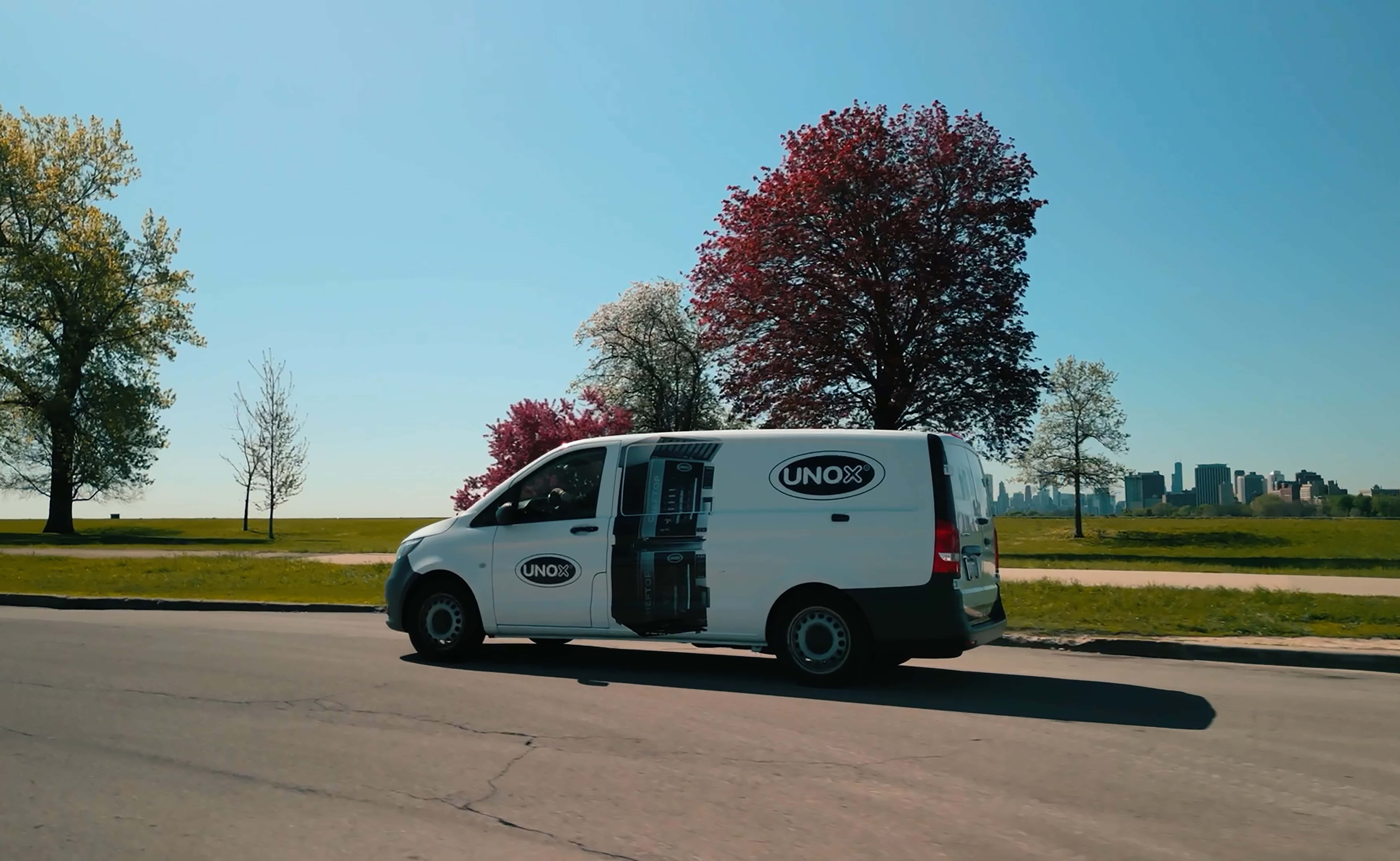 White van with Unox logo and images printed on drives down a road surrounded by green fields and trees.