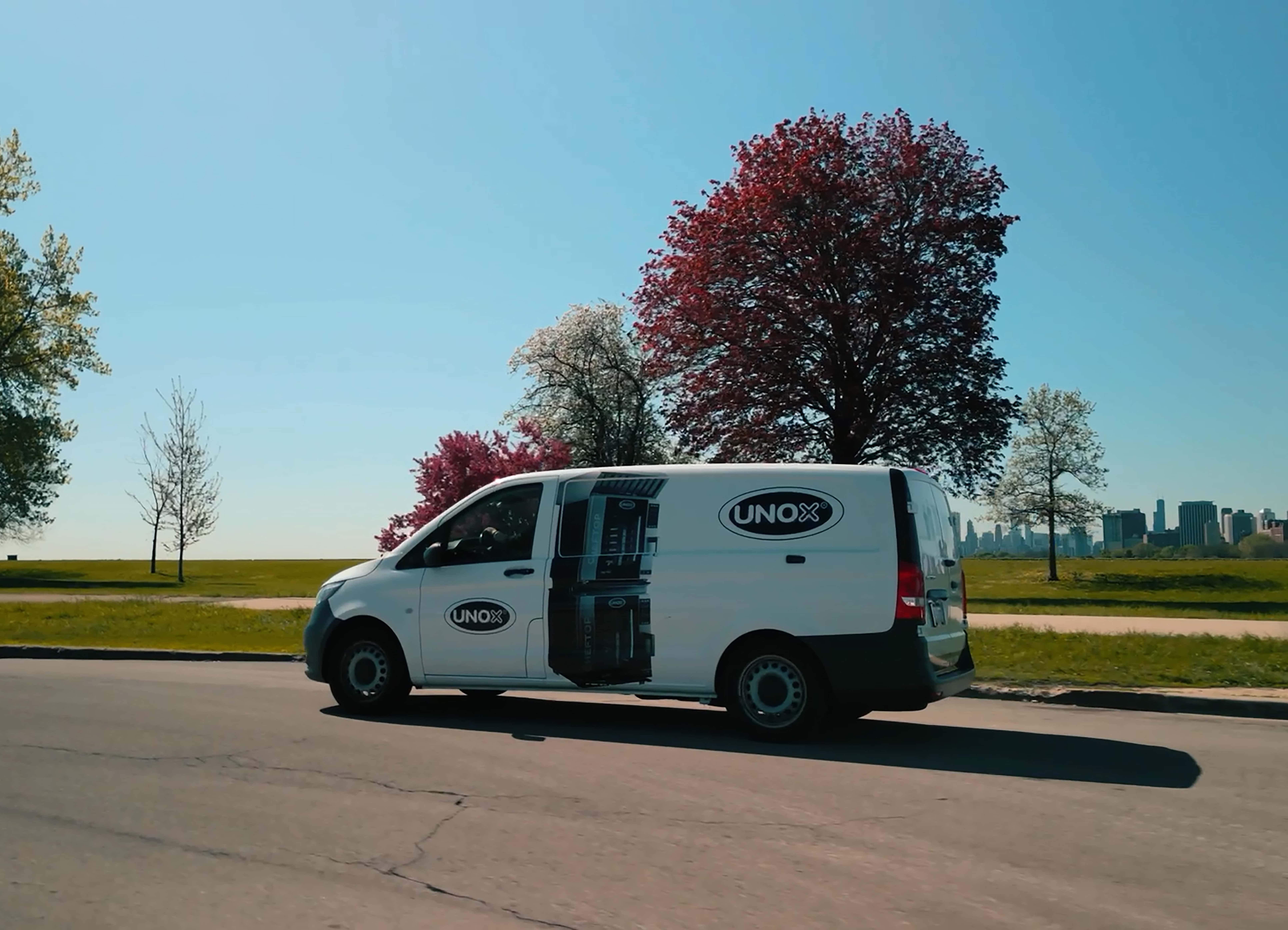 White van with Unox logo and images printed on drives down a road surrounded by green fields and trees.