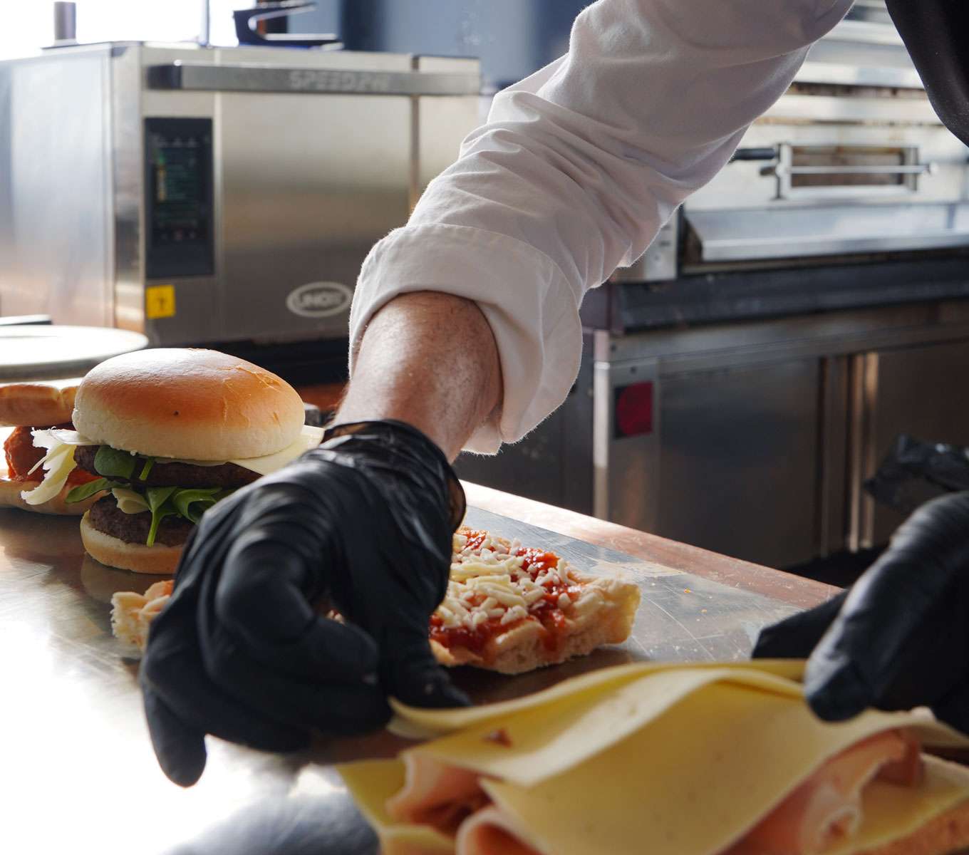 An operator prepares sandwiches and a pizza on the counter of a professional kitchen, with a small commercial speed oven in the background