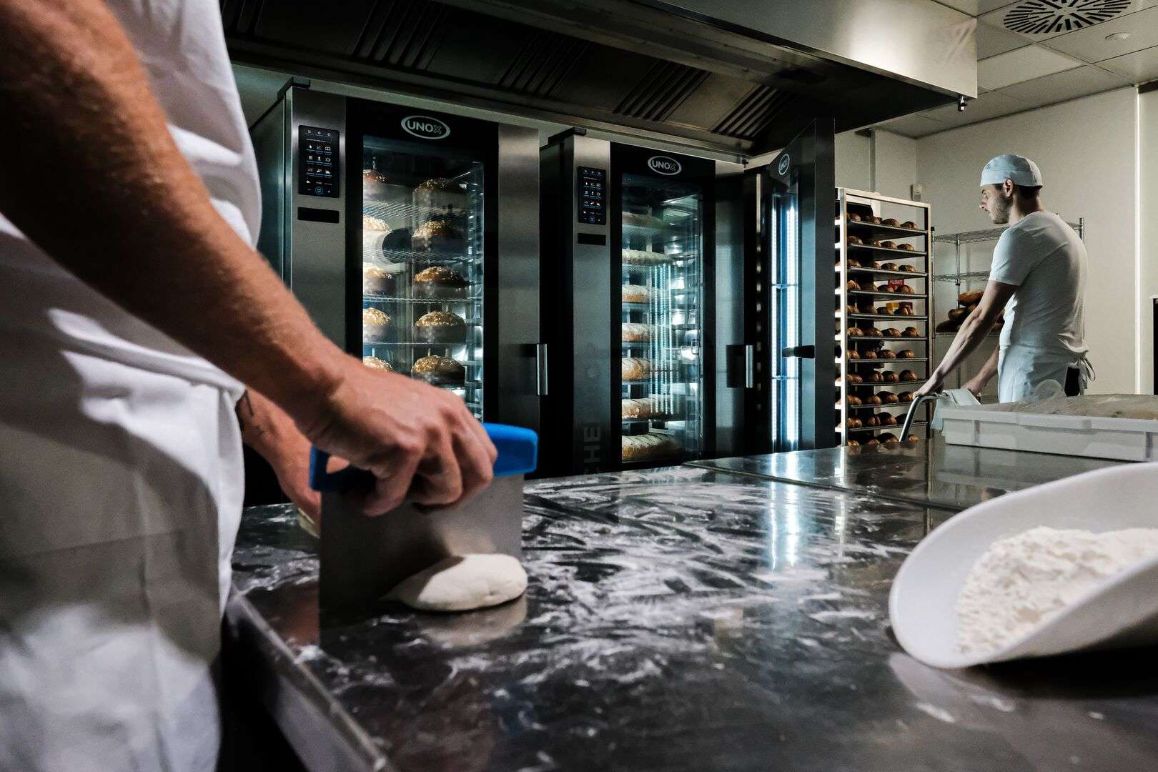 One operator prepares the dough while another inserts a cart into a Unox oven, while two other commercial ovens bake panettone