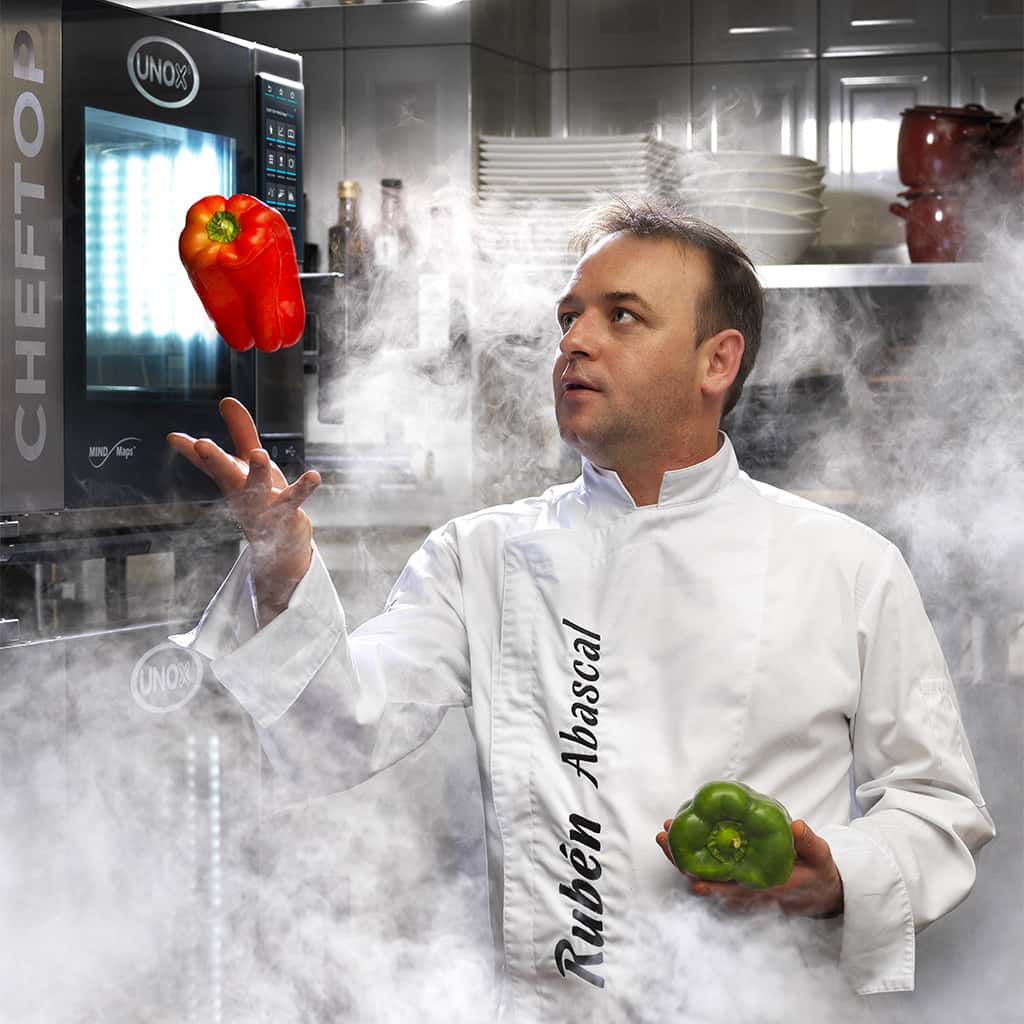 Male chef in white jacket stood in kitchen surrounded by smoke. He is throwing a red pepper in the air, stood next to an Unox combi oven.