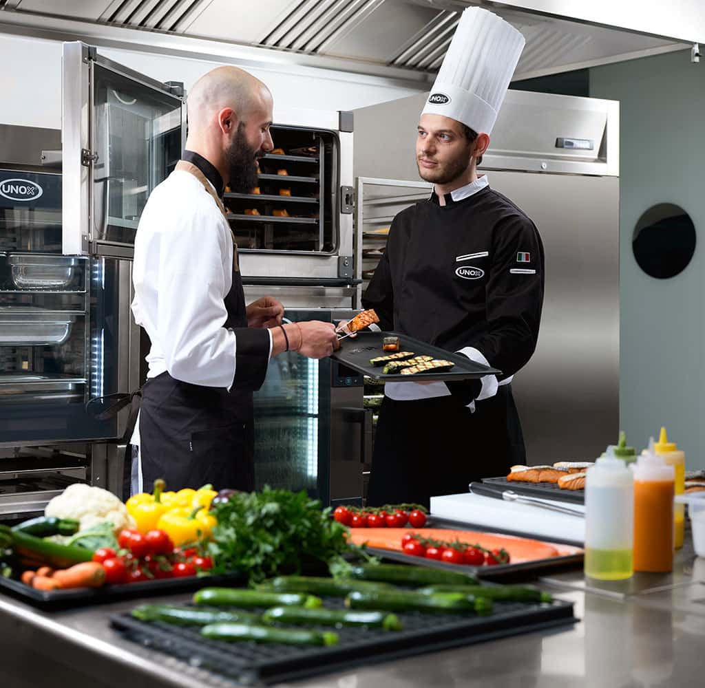 Two male chefs in kitchen in front of open combi oven, one holding tray of grilled vegetables and salmon, one picking up salmon from tray with tongs.