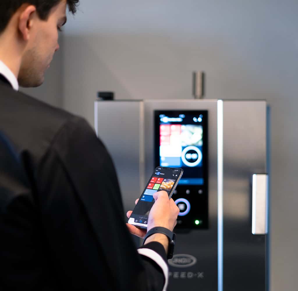 Man in black chef jacket holding a smart-phone pressing some buttons, facing towards an unox speed-x speed oven, controlling the oven with his phone.
