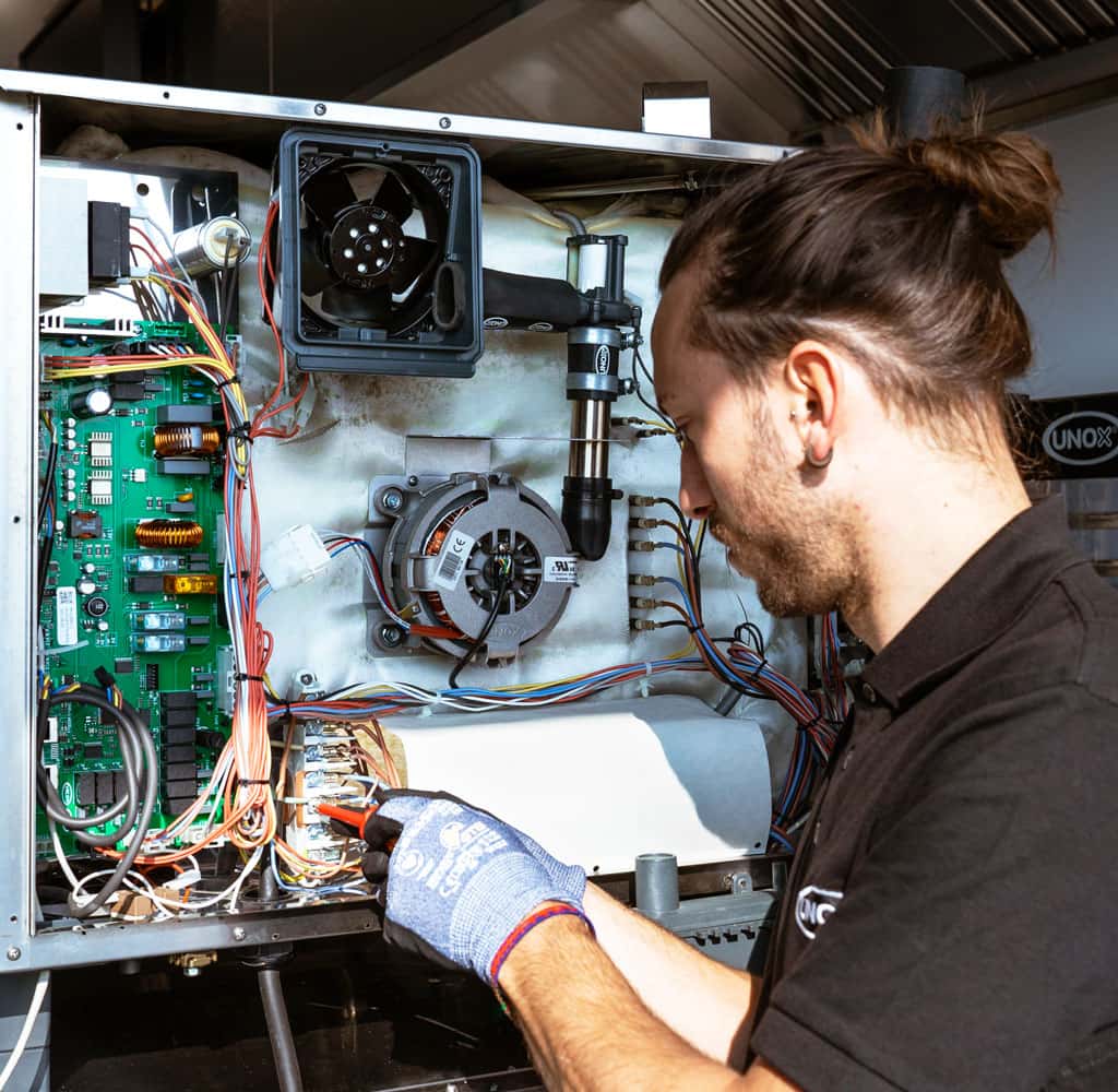 Male engineer wearing gloves, using a red screwdriver on the inside of a combi oven which has been disassembled, wires and motherboards can be seen.