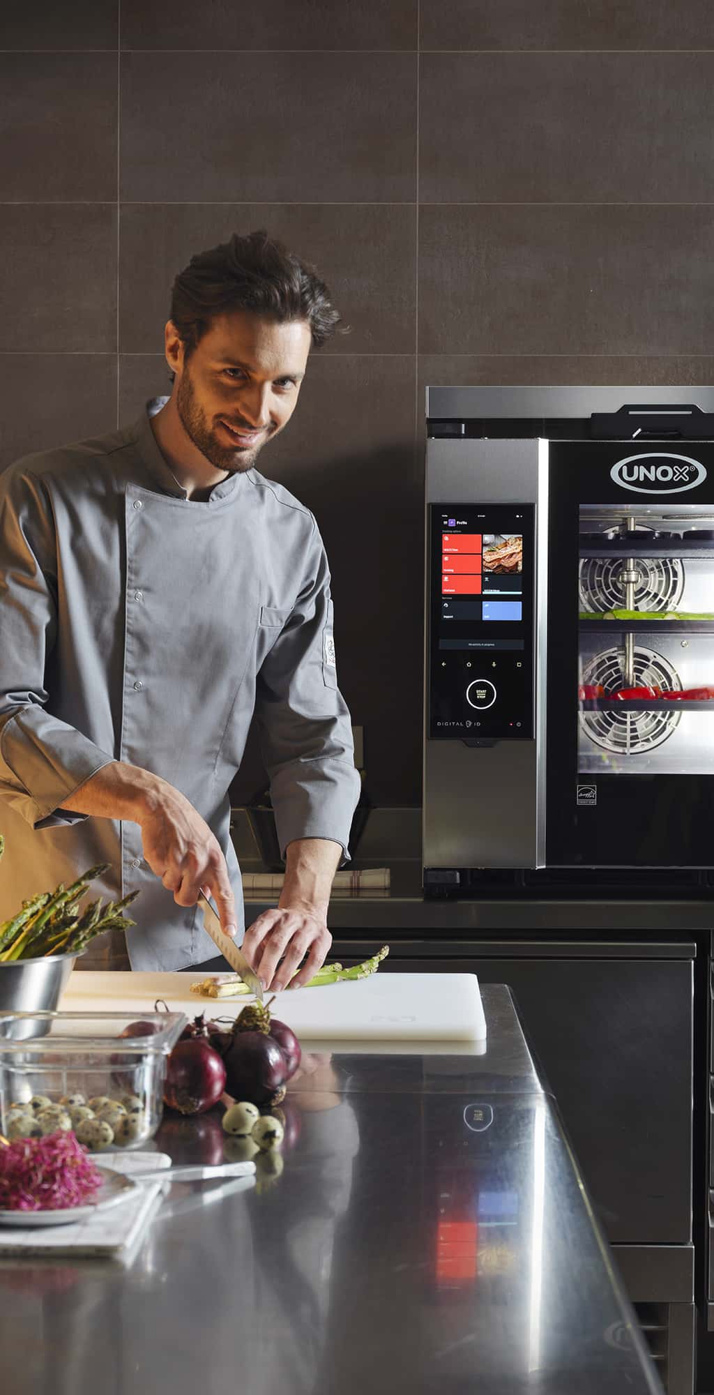 Smiling male chef looking towards the camera, chopping asparagus with a knife, stood in front of an Unox Cheftop-x combi oven.