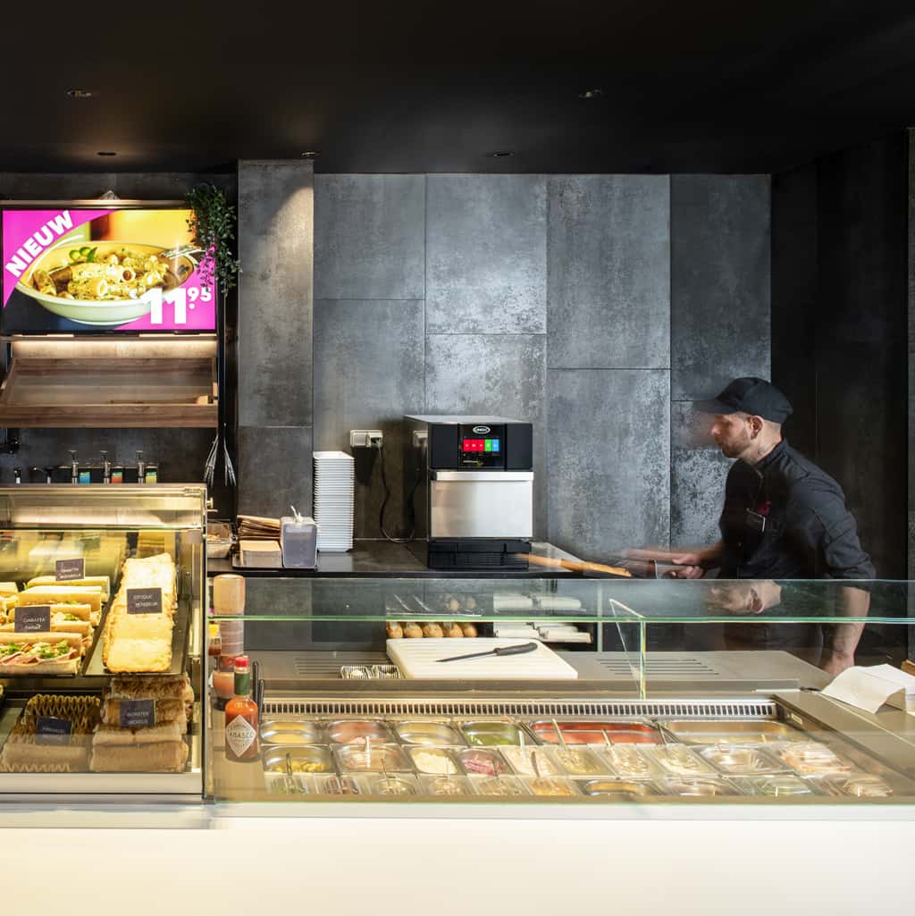 QSR male operator looking at an Unox speed oven behind a counter with wraps, sandwiches, and salads.