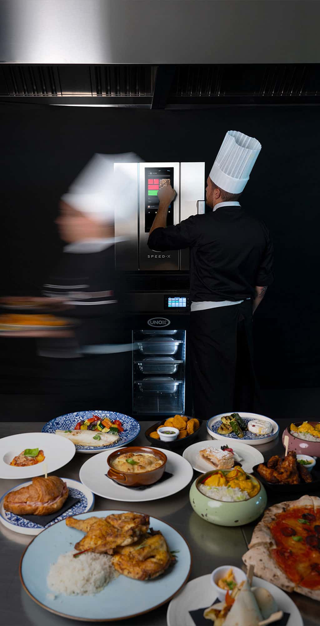 Kitchen counter full of dishes of food, a chef touches display of an Unox speed oven stacked on top of an evereo filled with trays.