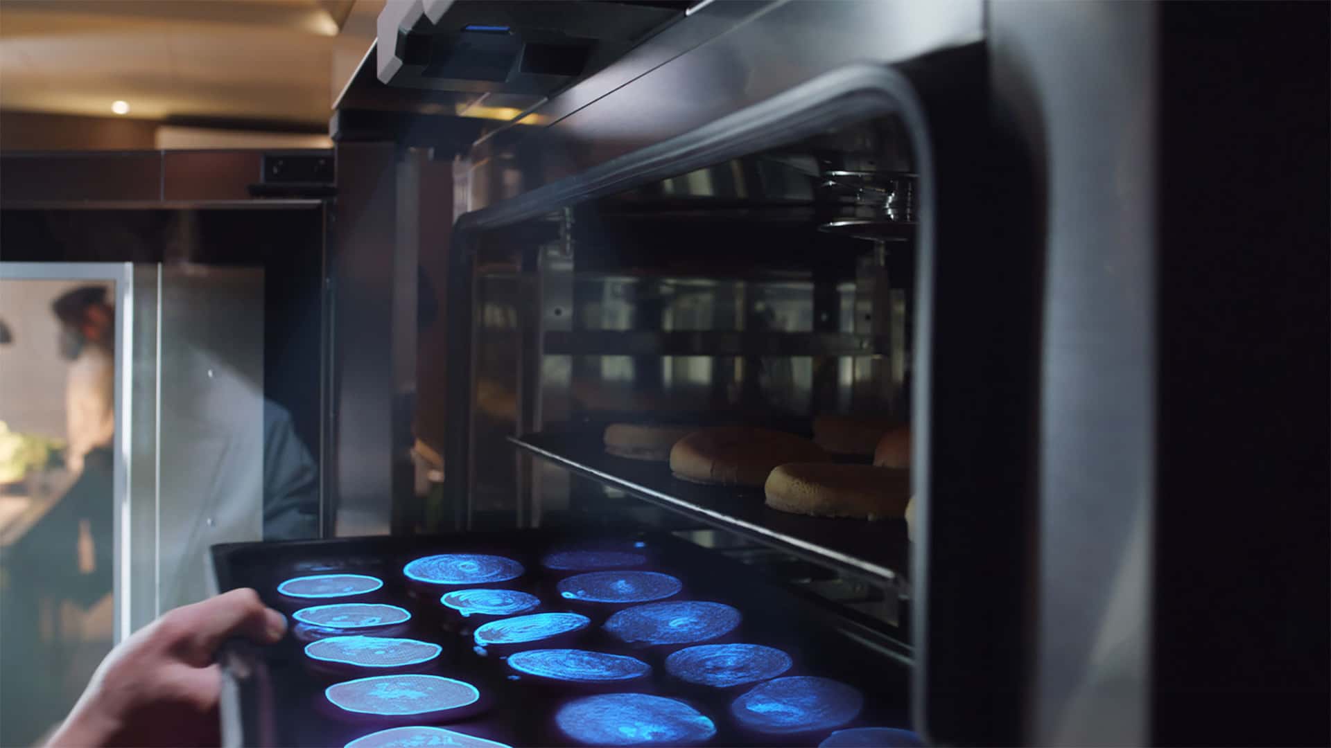 A hand puts a tray of food into an oven. A sensor at the top shines a blue light on the food to scan and identify what the food is.