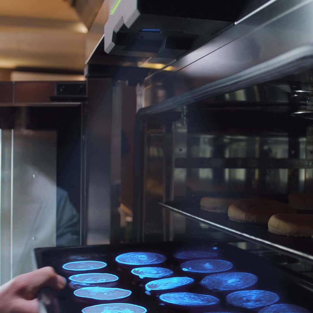 A hand puts a tray of food into an oven. A sensor at the top shines a blue light on the food to scan and identify what the food is.