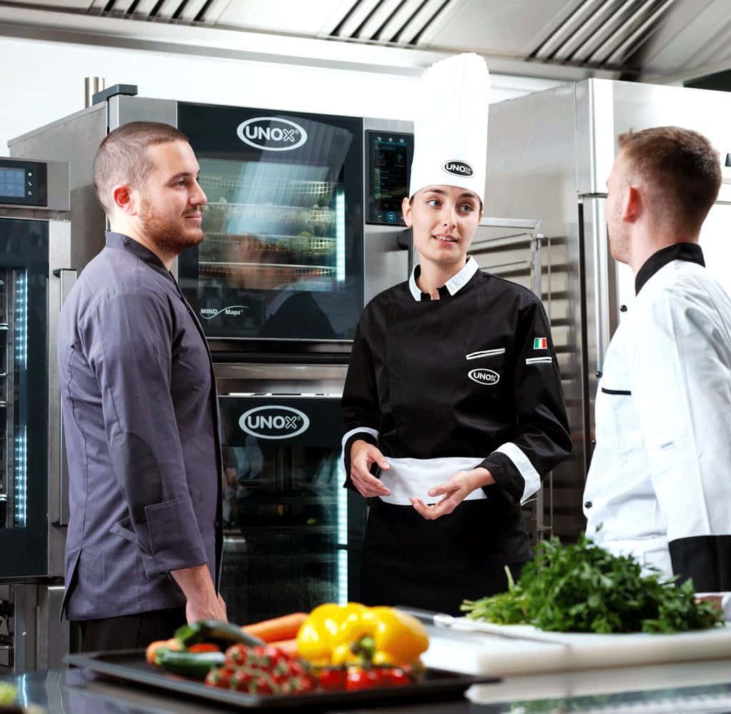 Two male chefs and a female chef stood talking in a kitchen in front of an Unox combi oven, counter with vegetables and herbs on.