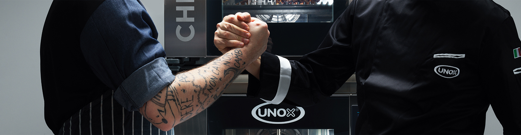 A male chef and a man wearing Unox uniform holding hands in front of an Unox combi oven.