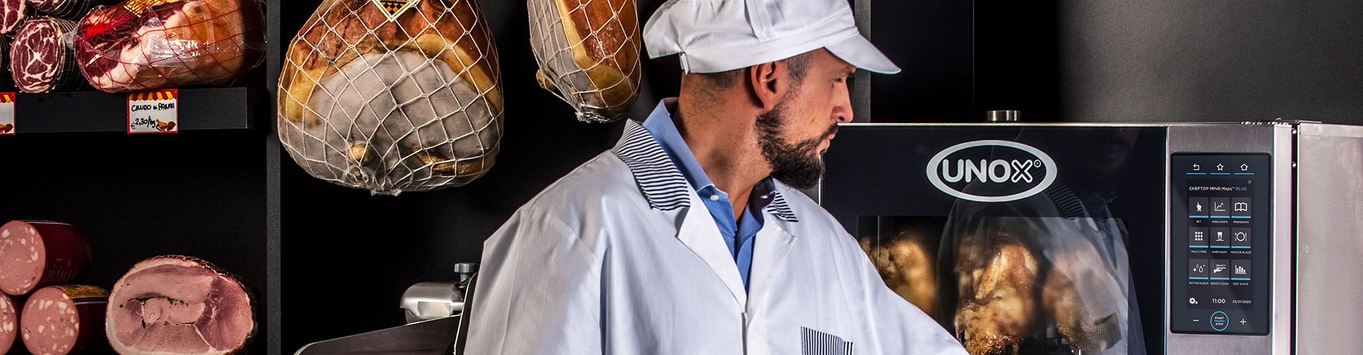 Man in a deli or butchers wearing a white hat, hams hang in the background, opening the door of an Unox combi oven filled with roasted chickens.