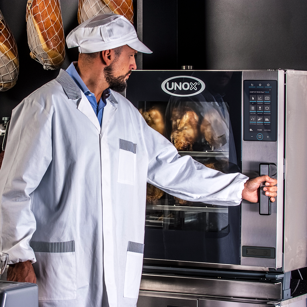 Man in a deli or butchers wearing a white hat, hams hang in the background, opening the door of an Unox combi oven filled with roasted chickens.