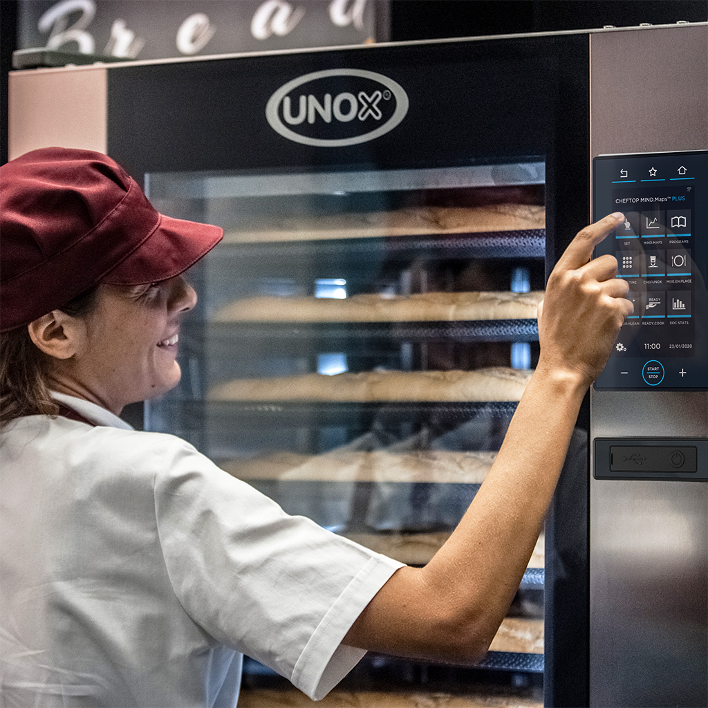 Woman in hat and uniform, pressing button on digital display panel on an Unox oven, bread baking inside the oven.