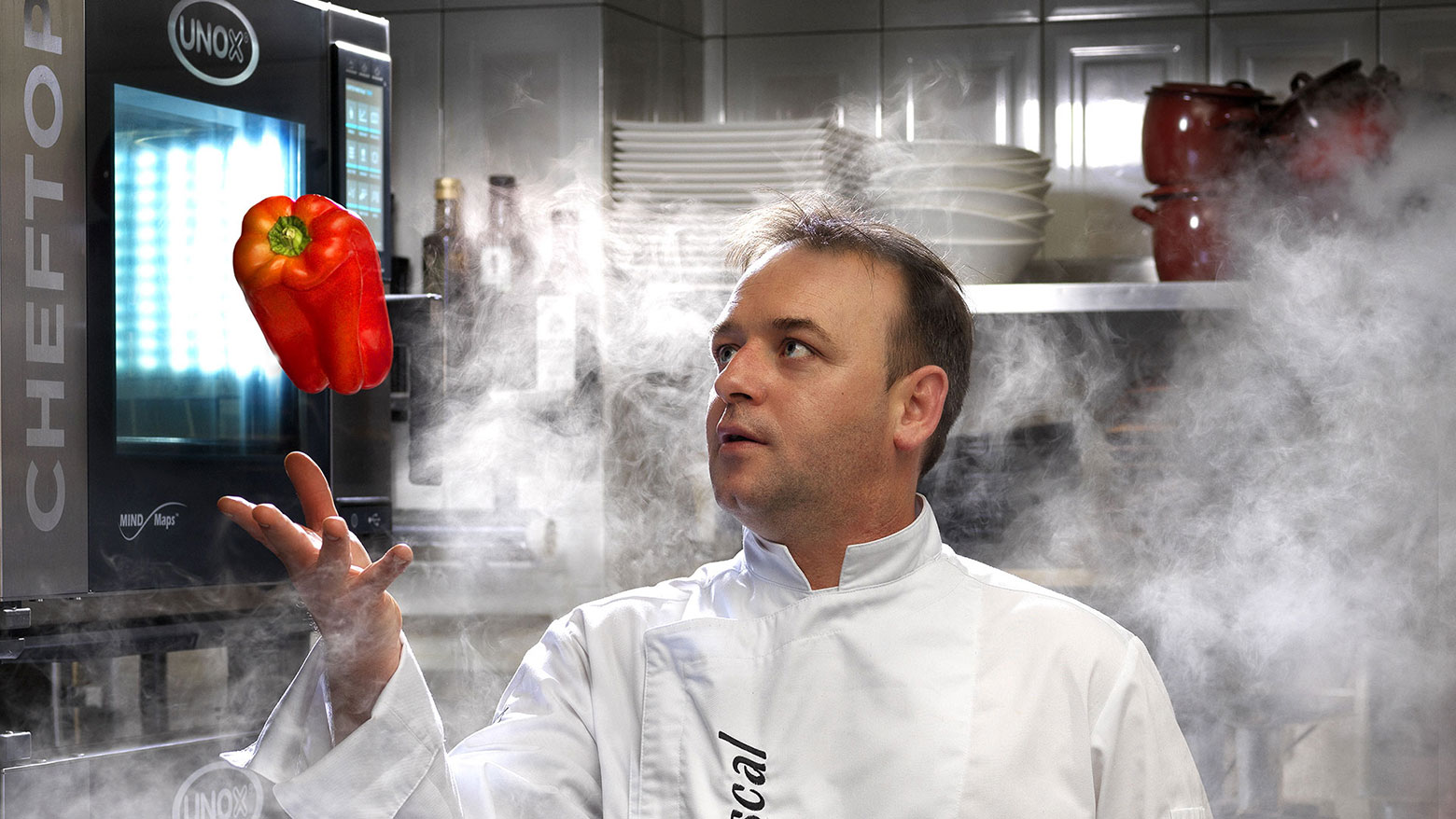 Male chef in white jacket stood in kitchen surrounded by smoke. He is throwing a red pepper in the air, stood next to an Unox combi oven.