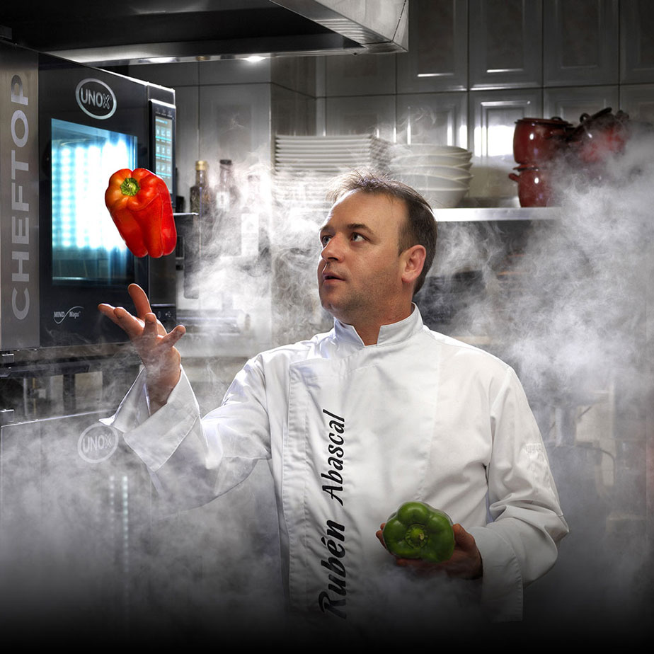 Male chef in white jacket stood in kitchen surrounded by smoke. He is throwing a red pepper in the air, stood next to an Unox combi oven.